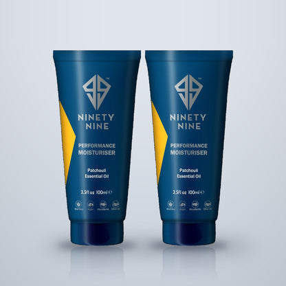 Two blue tubes of Ninety Nine Performance Moisturiser with Patchouli Essential Oil, displayed side-by-side on a light gray reflective surface.