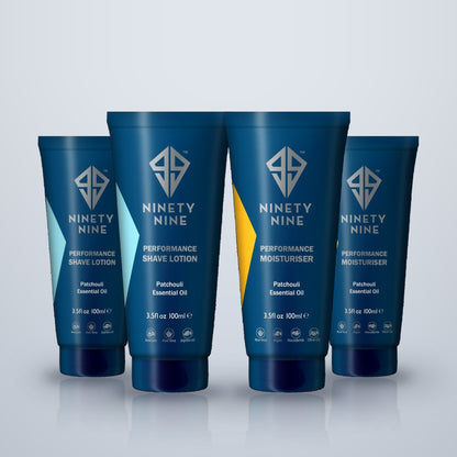 Four blue Ninety Nine personal care tubes standing upright on a light gray reflective surface. The tubes include Performance Shave Lotion, Performance Moisturiser, and two additional variants, all with white text and geometric logos.
